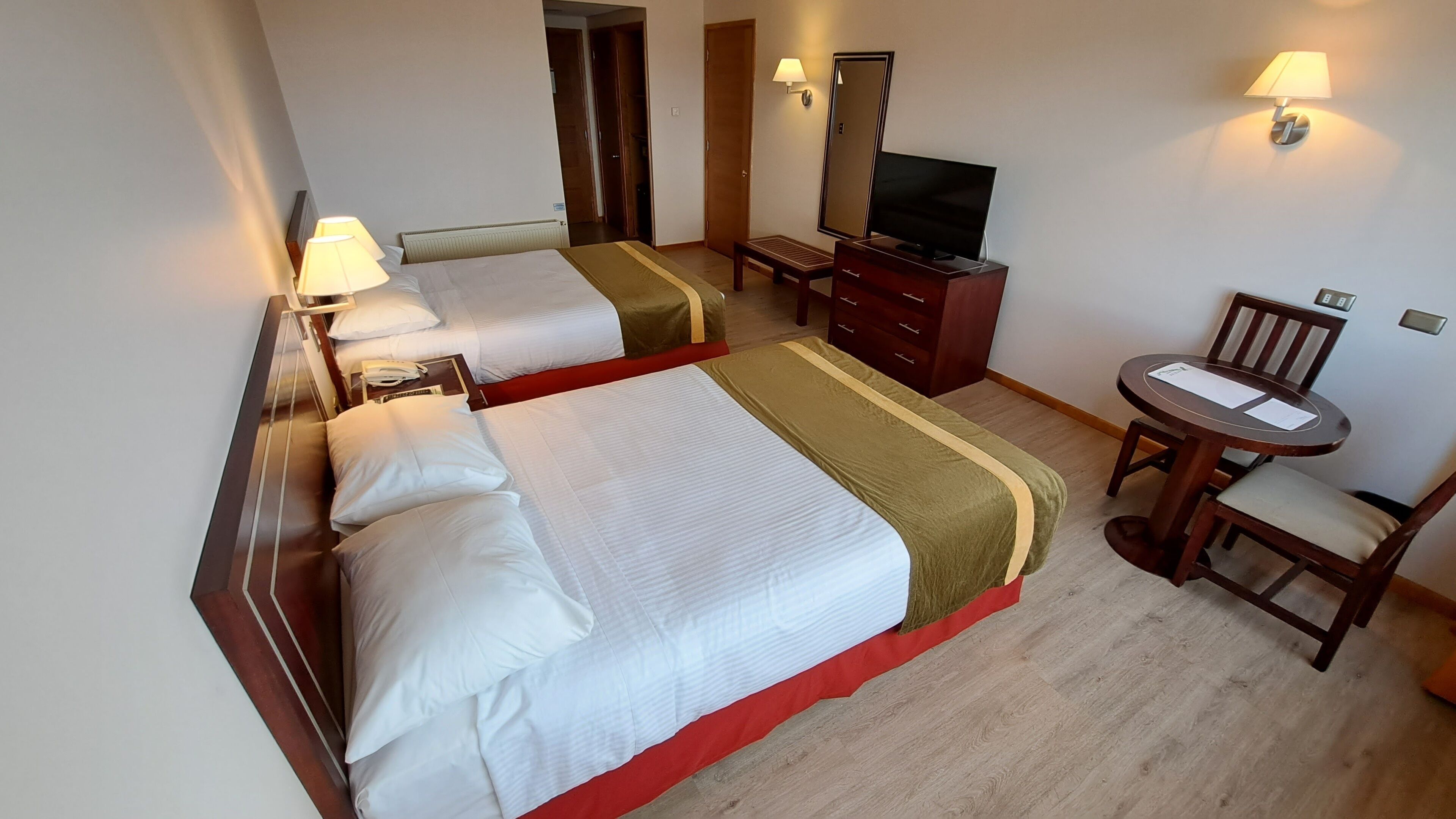 A hotel room with two beds, a desk, a chair, a TV, and a lamp.