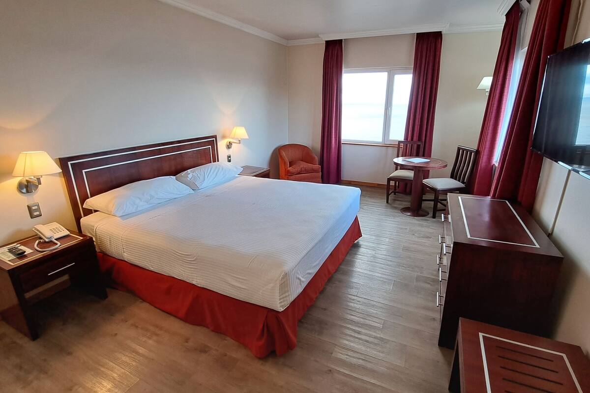 A hotel room with a large bed, bedside lamps, a desk, a chair, a TV, and a window with curtains.