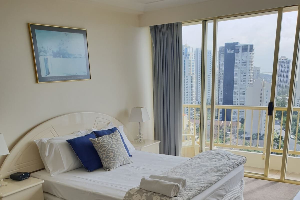 A hotel room with a large bed, a nightstand, a lamp, a framed picture on the wall, and a view of tall buildings through the window.