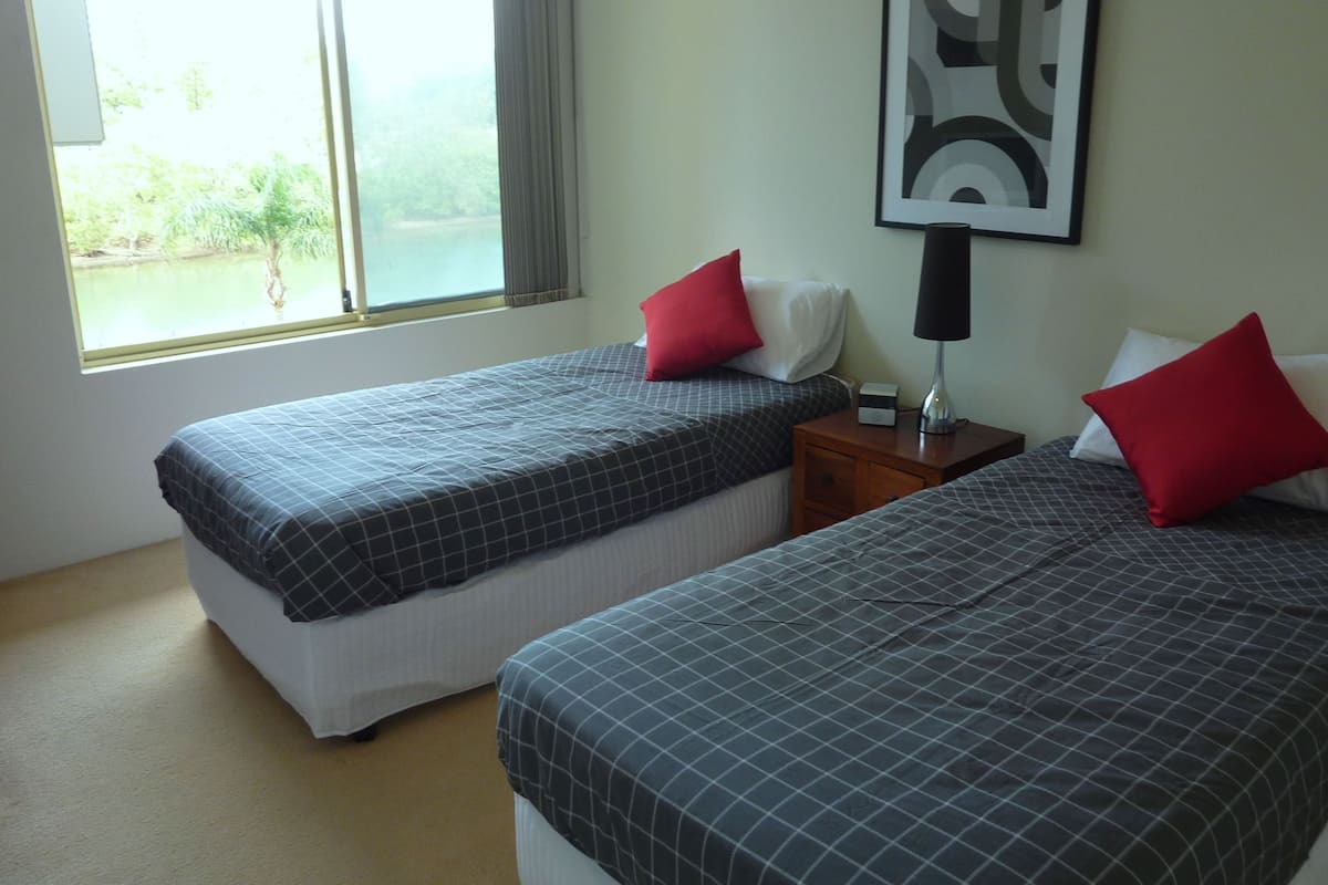 A hotel room with two beds, a nightstand with a lamp, a framed picture on the wall, and a window with a view of greenery.