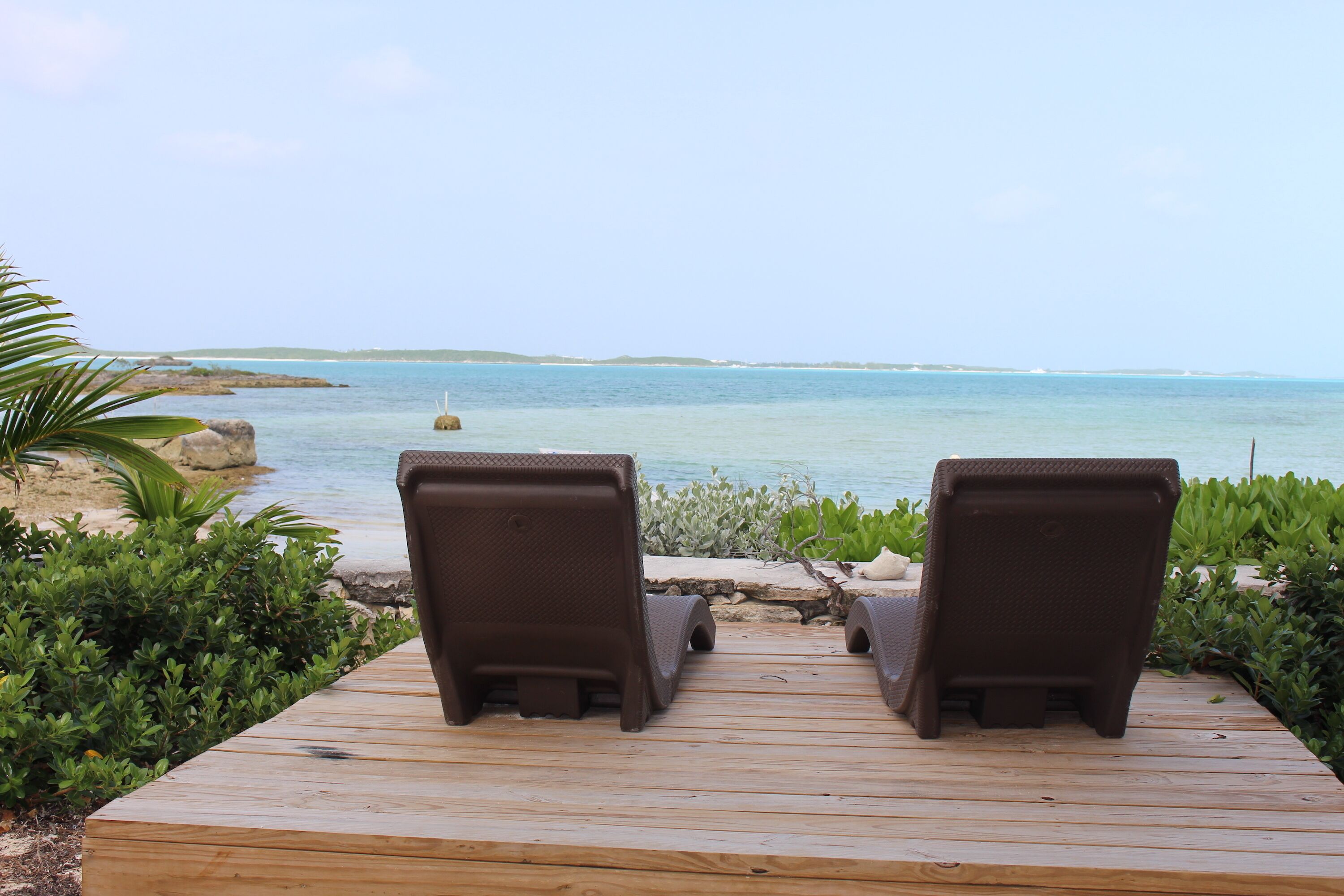 Two brown lounge chairs on a wooden deck overlooking a calm sea with distant land.
