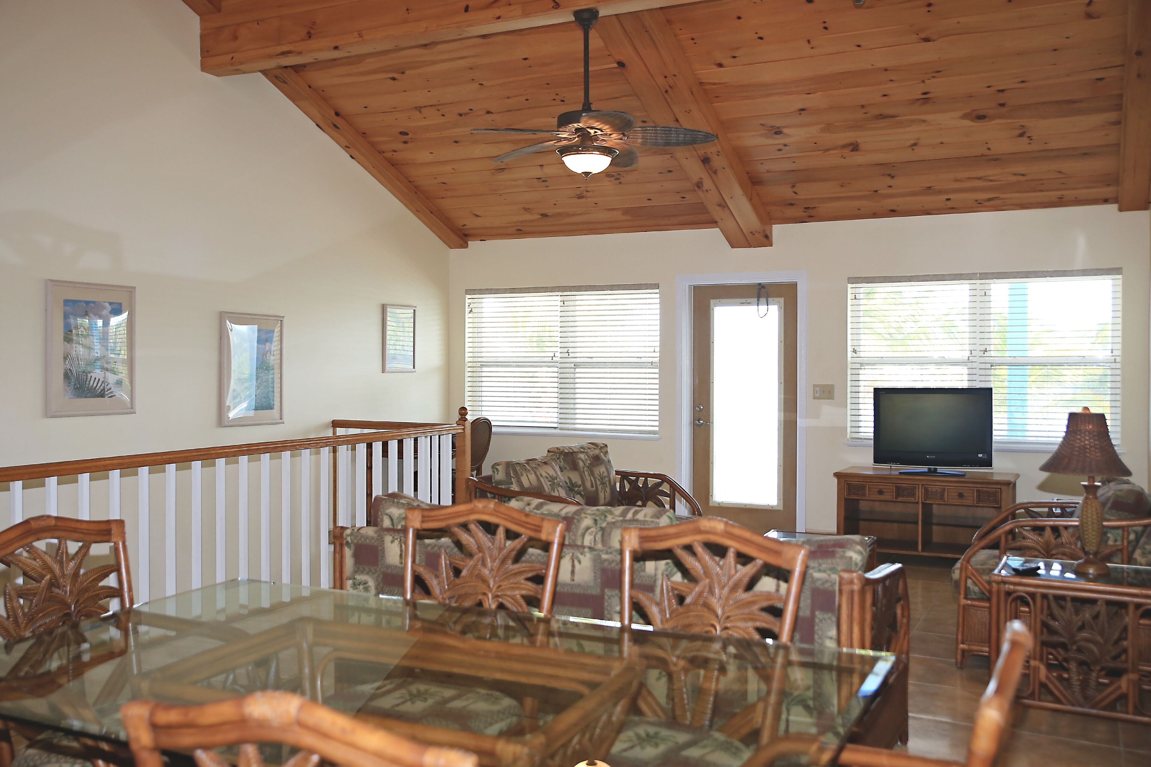 A spacious living room with a wooden ceiling, a glass dining table surrounded by chairs, a sofa, a television, and a lamp.