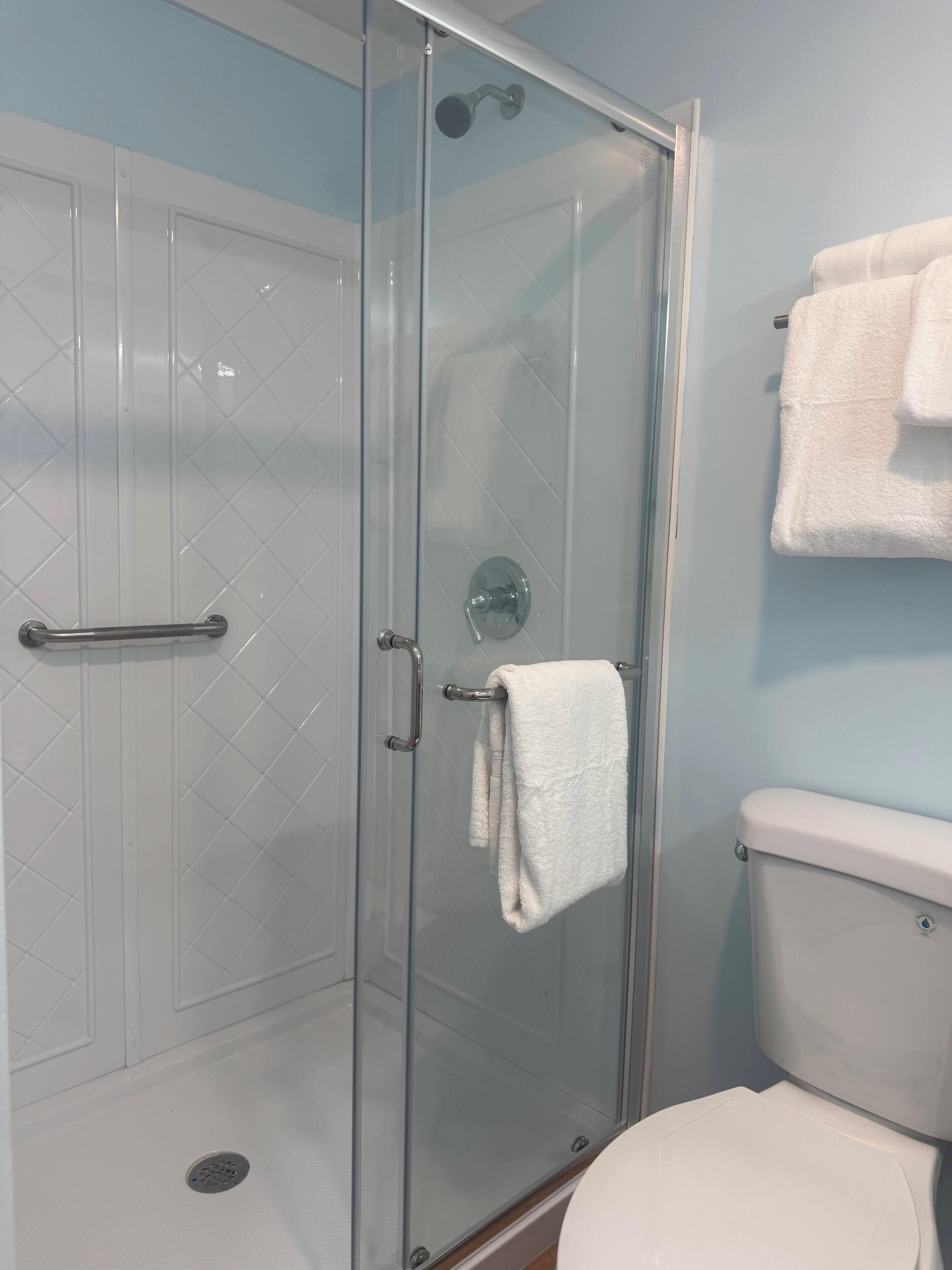 Een douche met glazen wanden, een witte betegelde muur, een wit toilet en een handdoekrek met handdoeken.