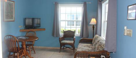 A room with a dining table, two chairs, a television, a sofa, a lamp, and a window with curtains.