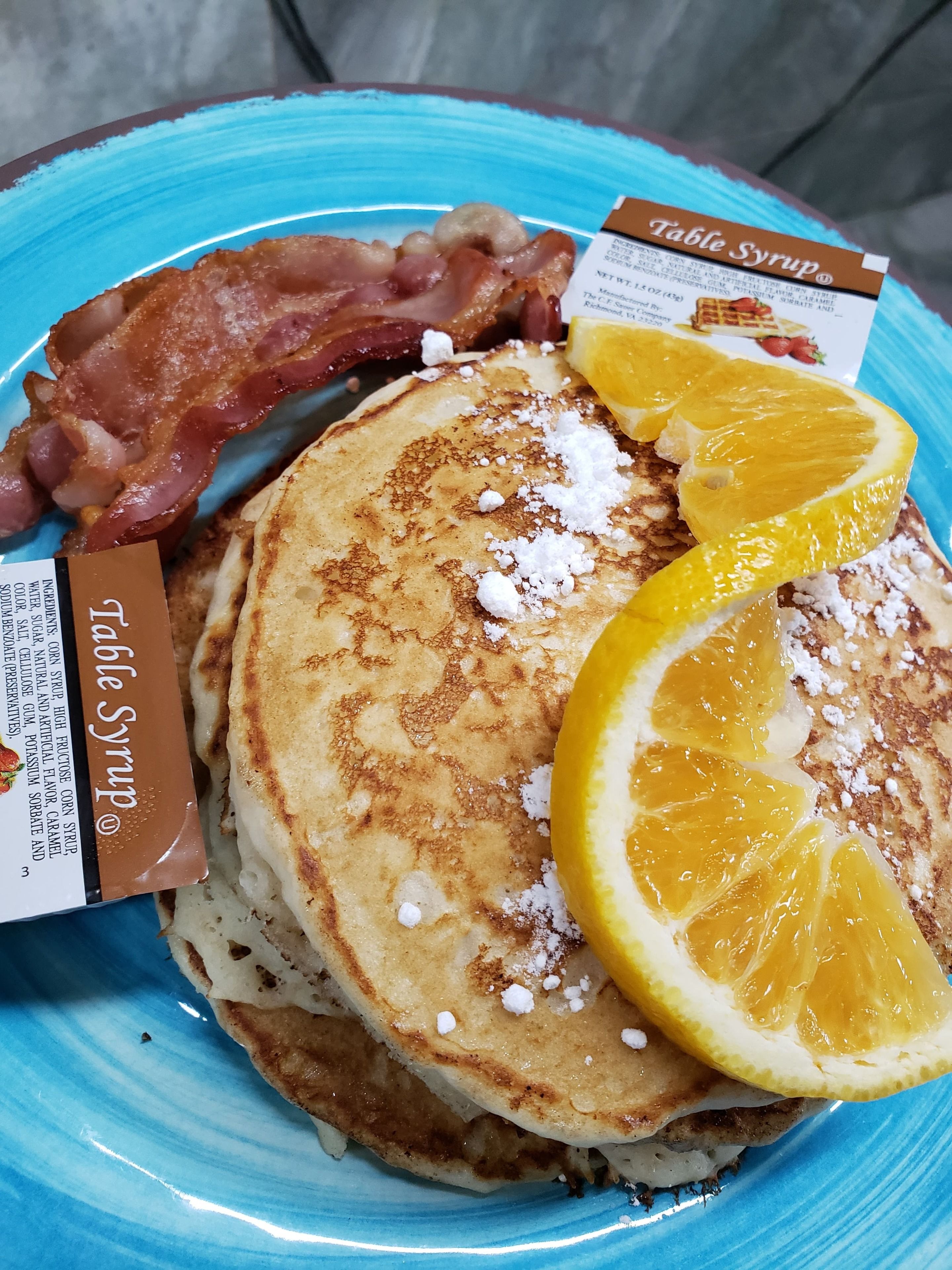 A plate with pancakes, bacon, and a slice of orange on a blue plate.