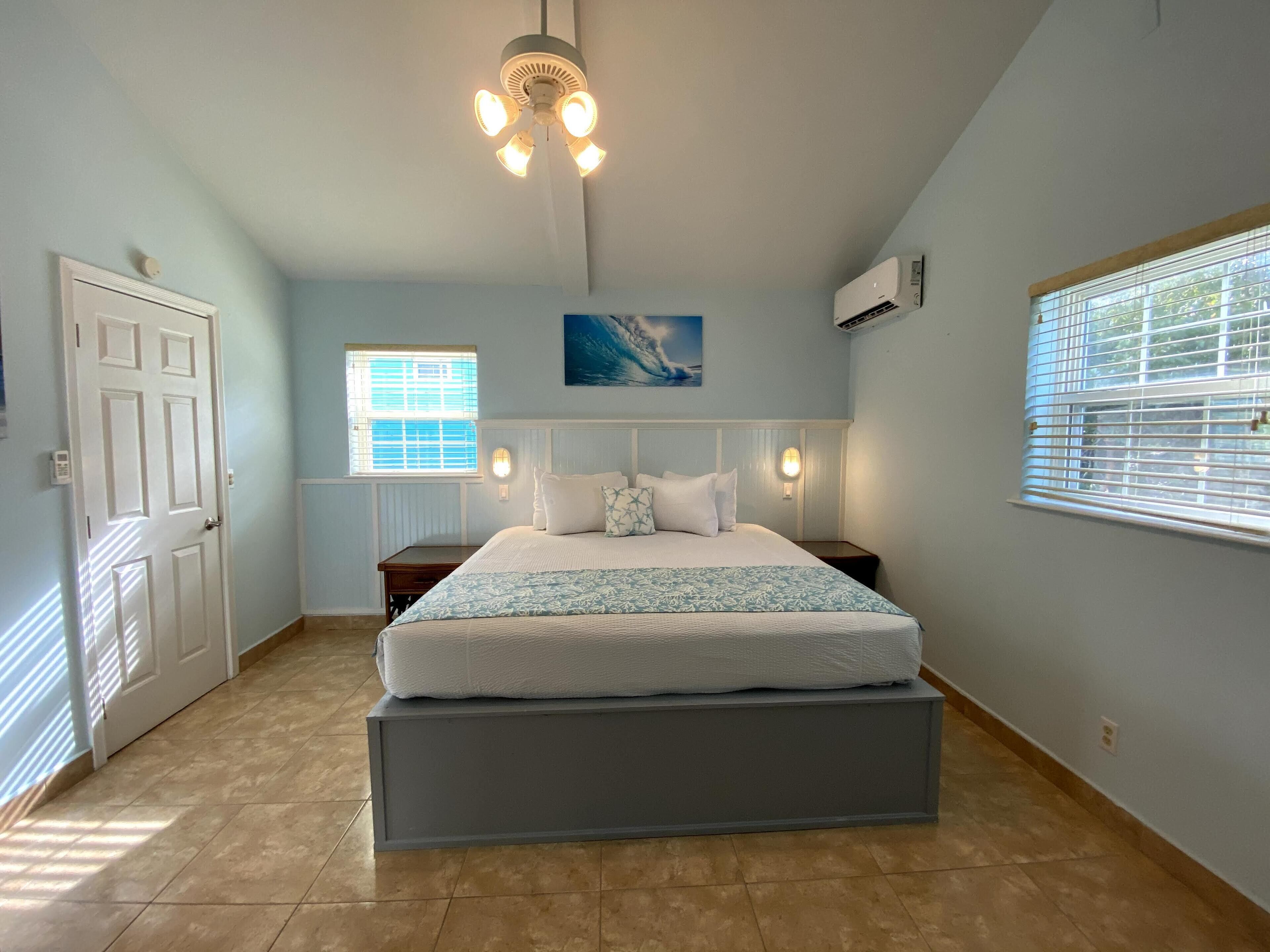 A bedroom with a bed, a ceiling fan, a window with blinds, an air conditioning unit, and a door.