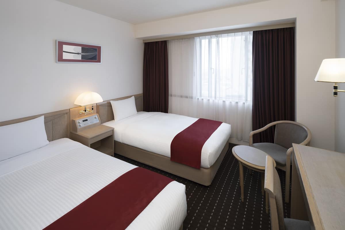 A hotel room with two beds, a desk, a chair, a lamp, and a window with curtains.