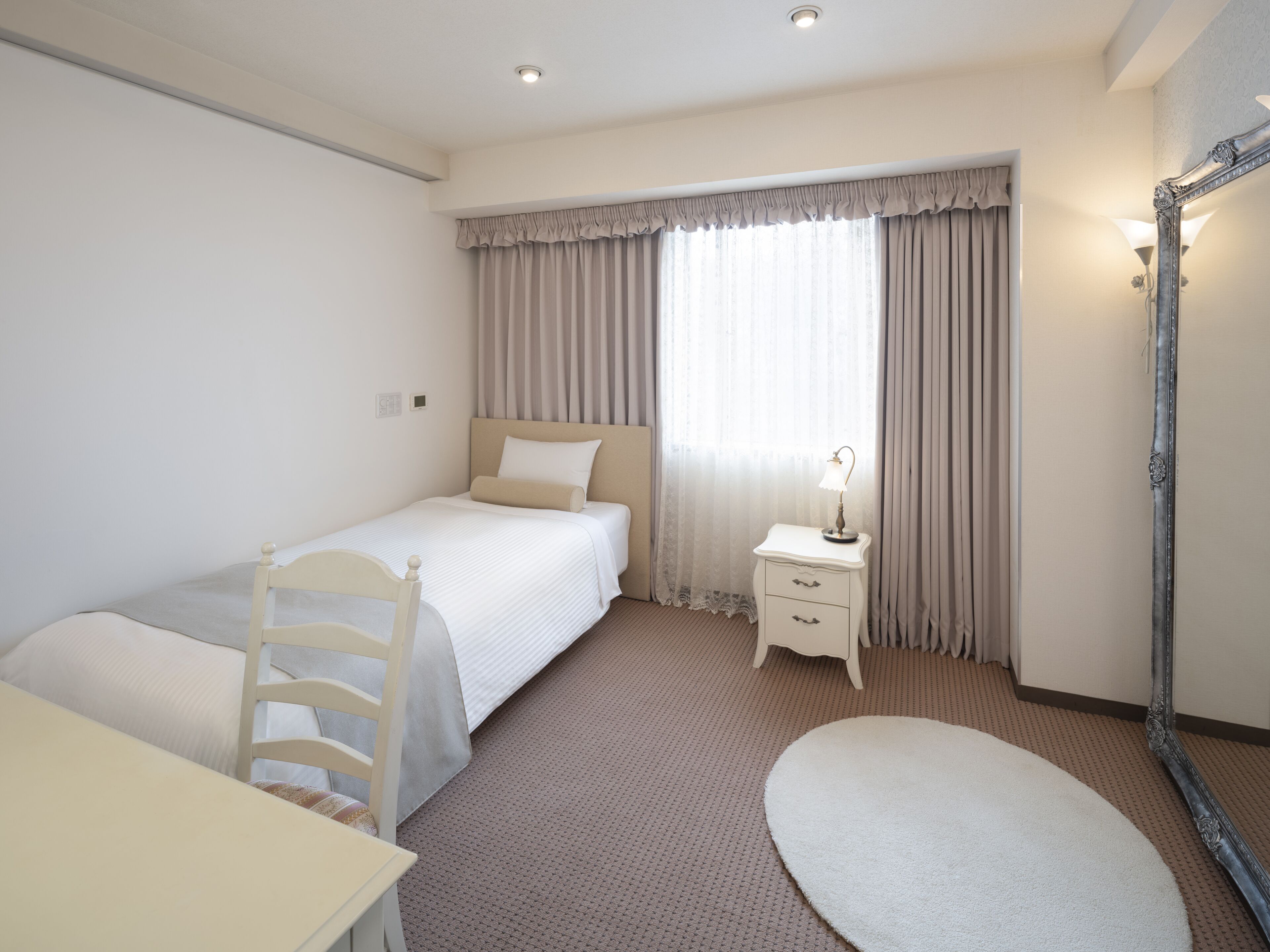 A hotel room with a single bed, a bedside table with a lamp, a chair, a desk, and a round rug.