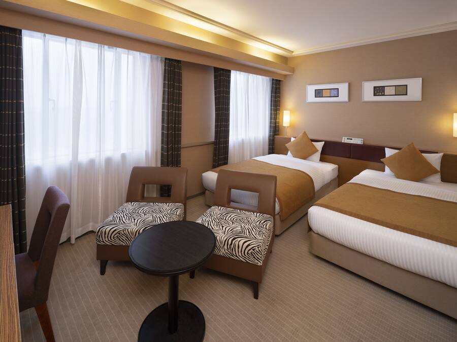 Une chambre d’hôtel avec deux lits, une petite table, deux chaises et une grande fenêtre avec des rideaux.