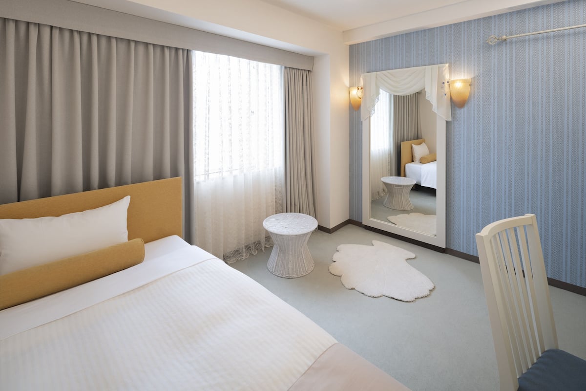 A hotel room with a large bed, a round side table, a chair, a mirror, and a view of another room.