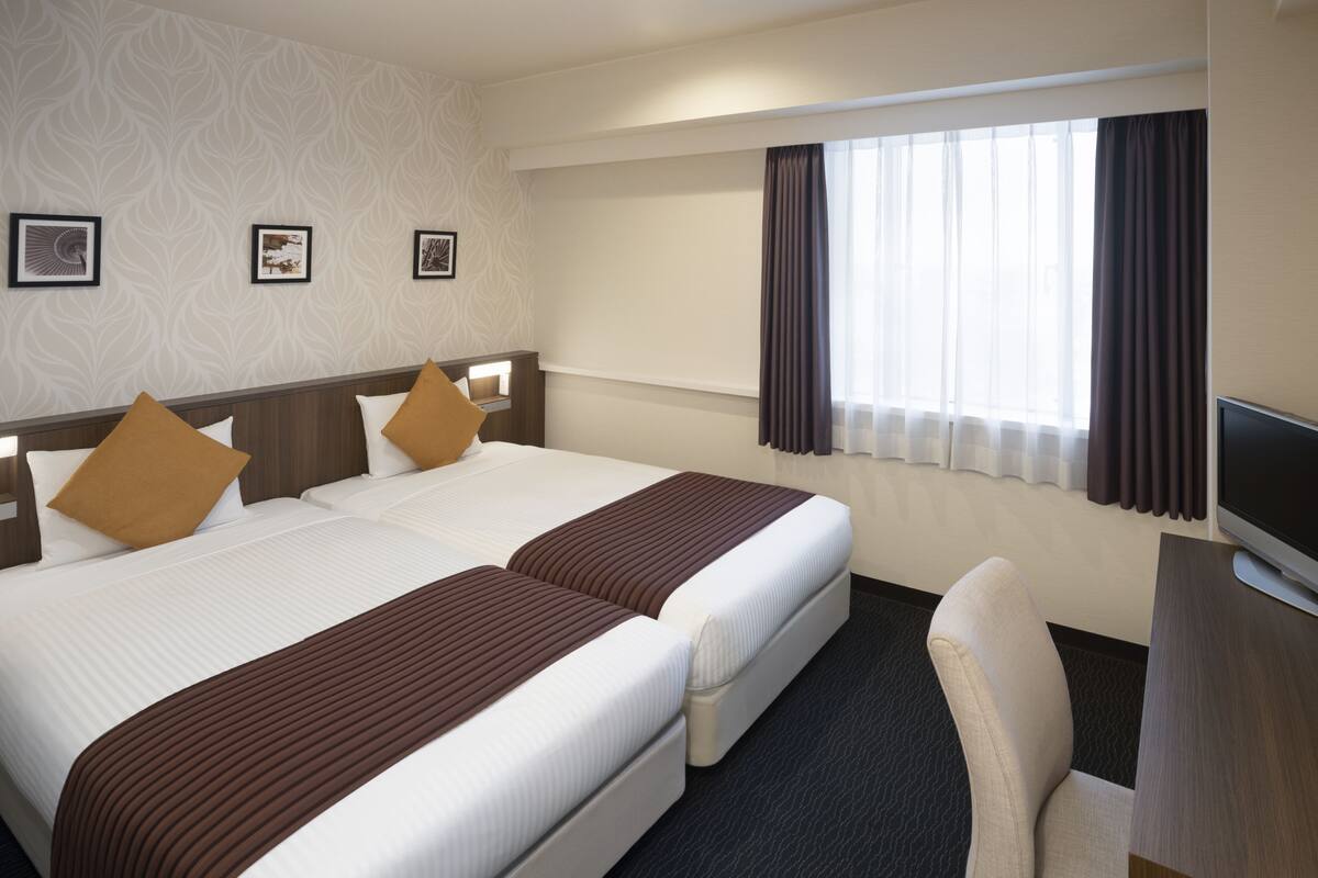 A hotel room with two beds, a desk, a chair, and a TV.