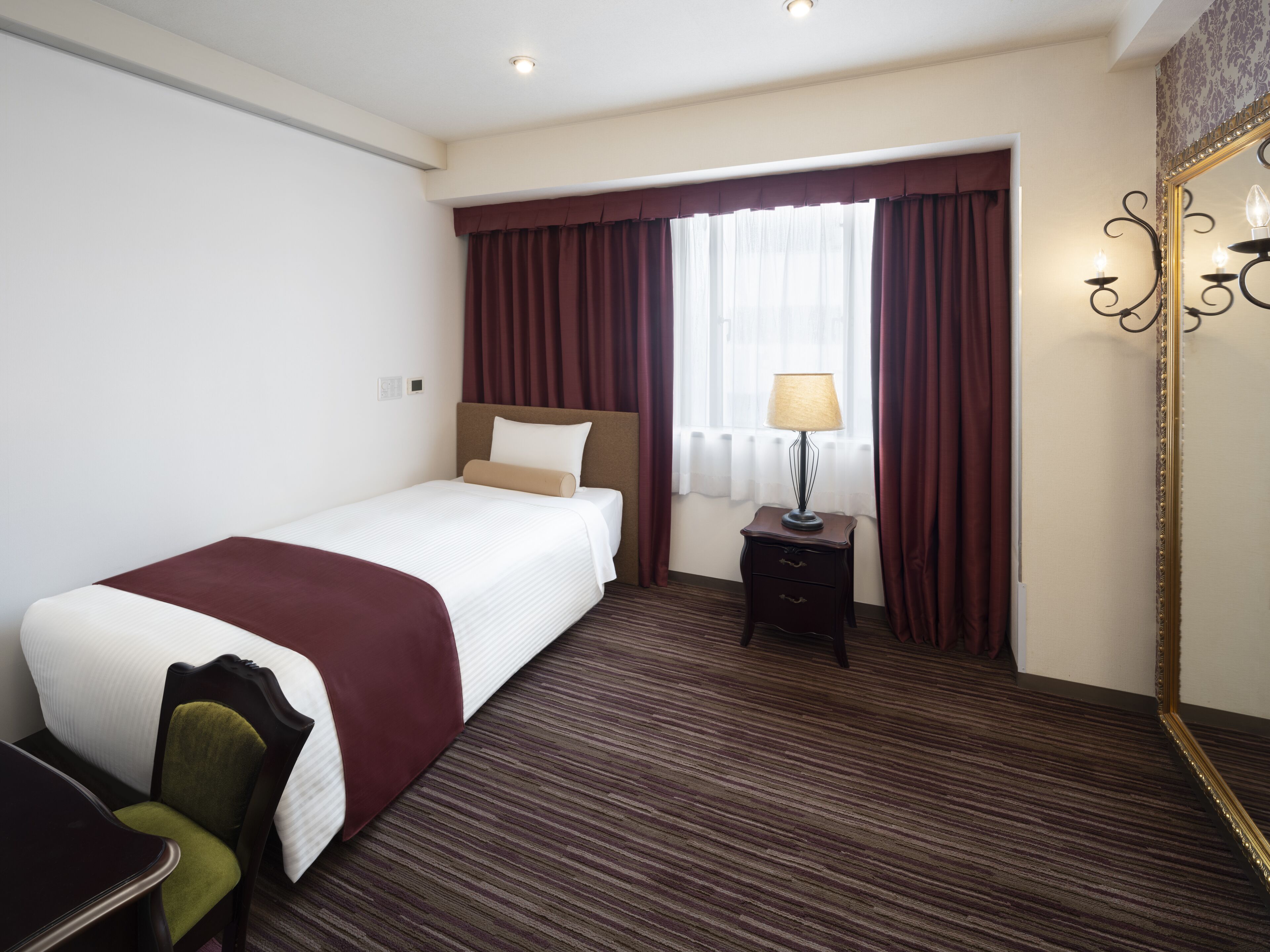 A hotel room with a bed, a nightstand with a lamp, a chair, and a large mirror.