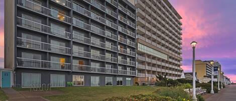A multi-story hotel with balconies, a paved walkway, and a landscaped area with bushes.
