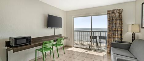 A room with a TV, a microwave, two green chairs, a grey sofa, a balcony with a view, and a table with a lamp.