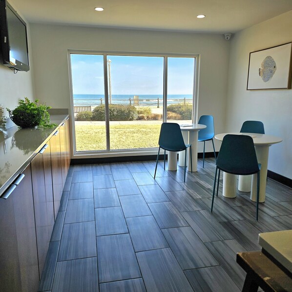 A modern room with a large sliding glass door leading to a balcony with a view of the beach and ocean. There is a small table with blue chairs and a potted plant on the left side.