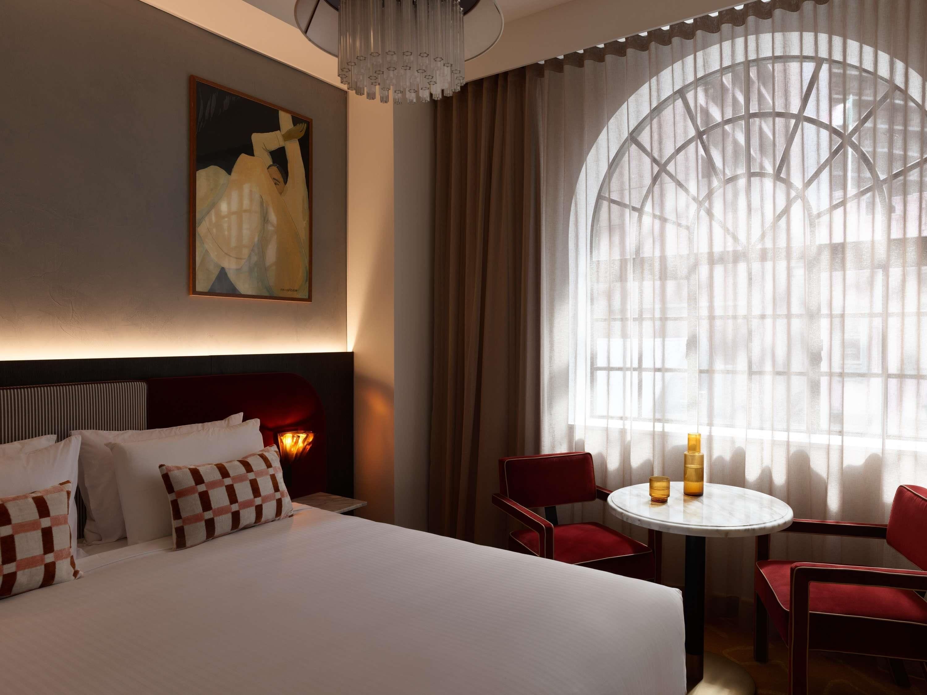 A hotel room with a large bed, a round table with chairs, a painting on the wall, and a large window with curtains.