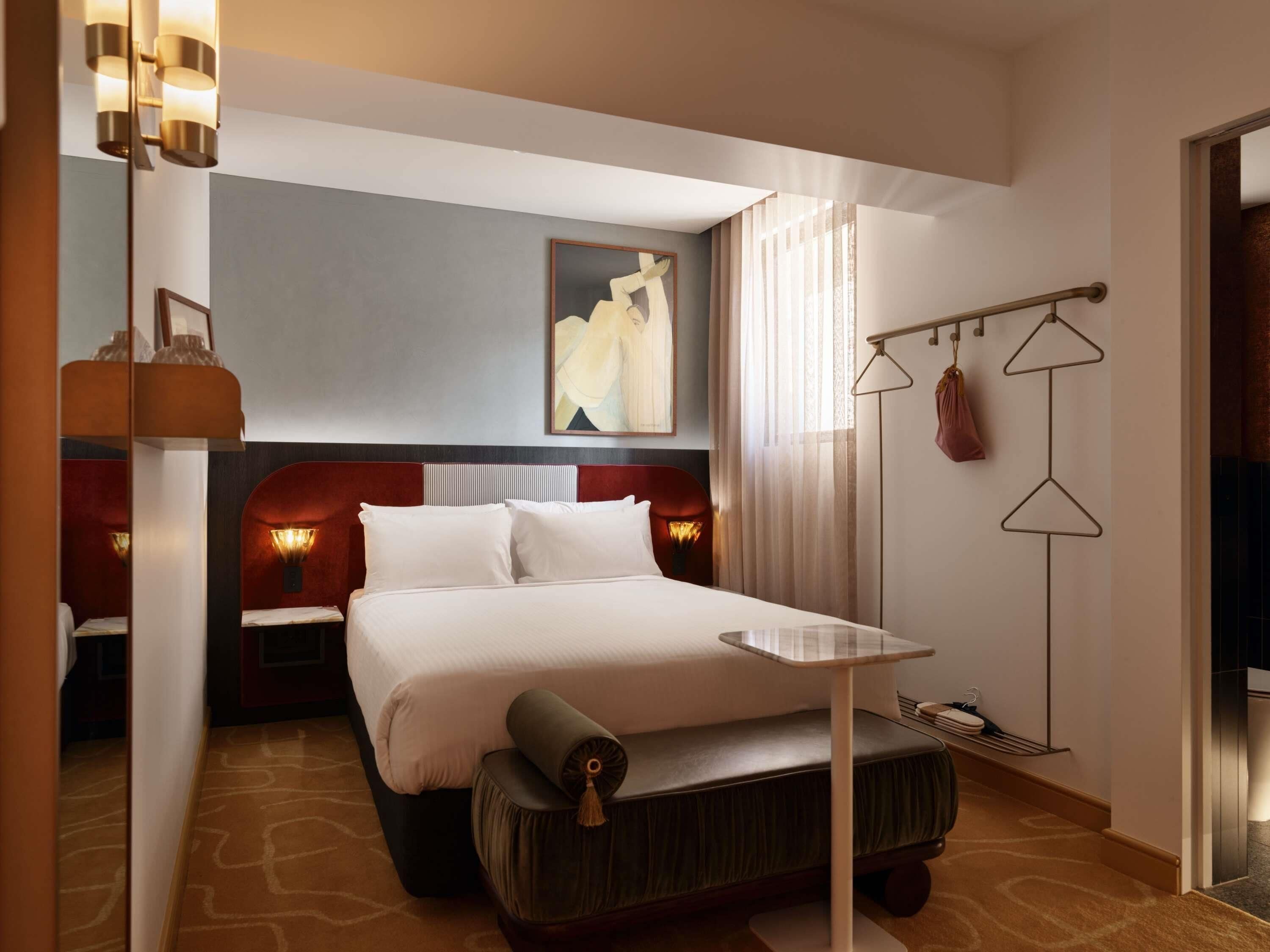 A hotel room with a bed, bedside tables, a small table, a chair, a floor lamp, and a painting on the wall.