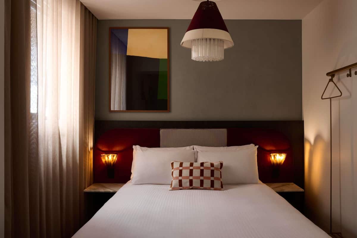 A hotel room with a large bed, two bedside lamps, a chandelier, and a painting on the wall.