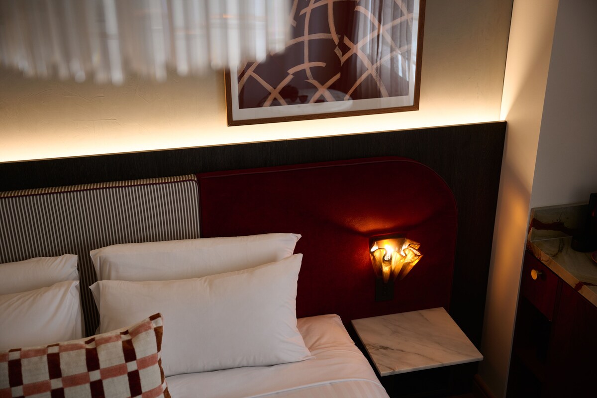 Quarto de hotel com cama, almofadas, mesa de cabeceira e luminária fixada na parede.
