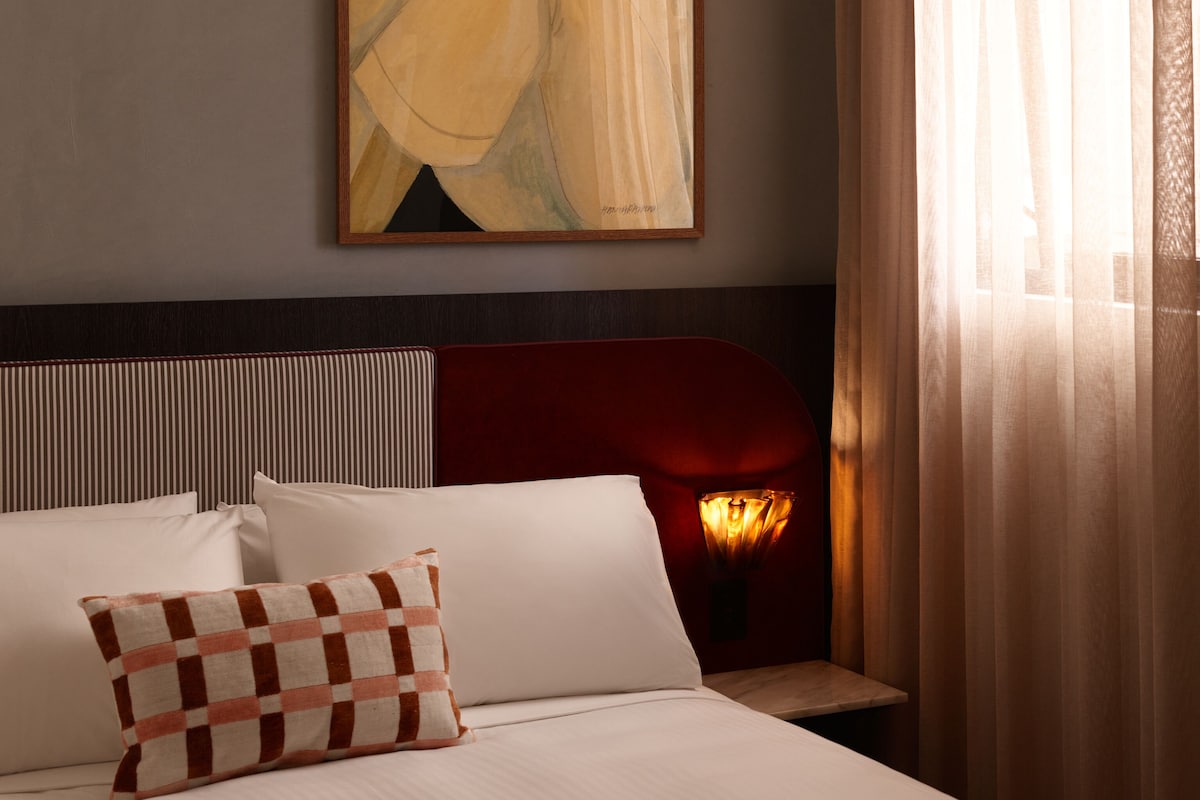 Quarto de hotel com cama, cabeceira, um quadro e janela com cortinas.