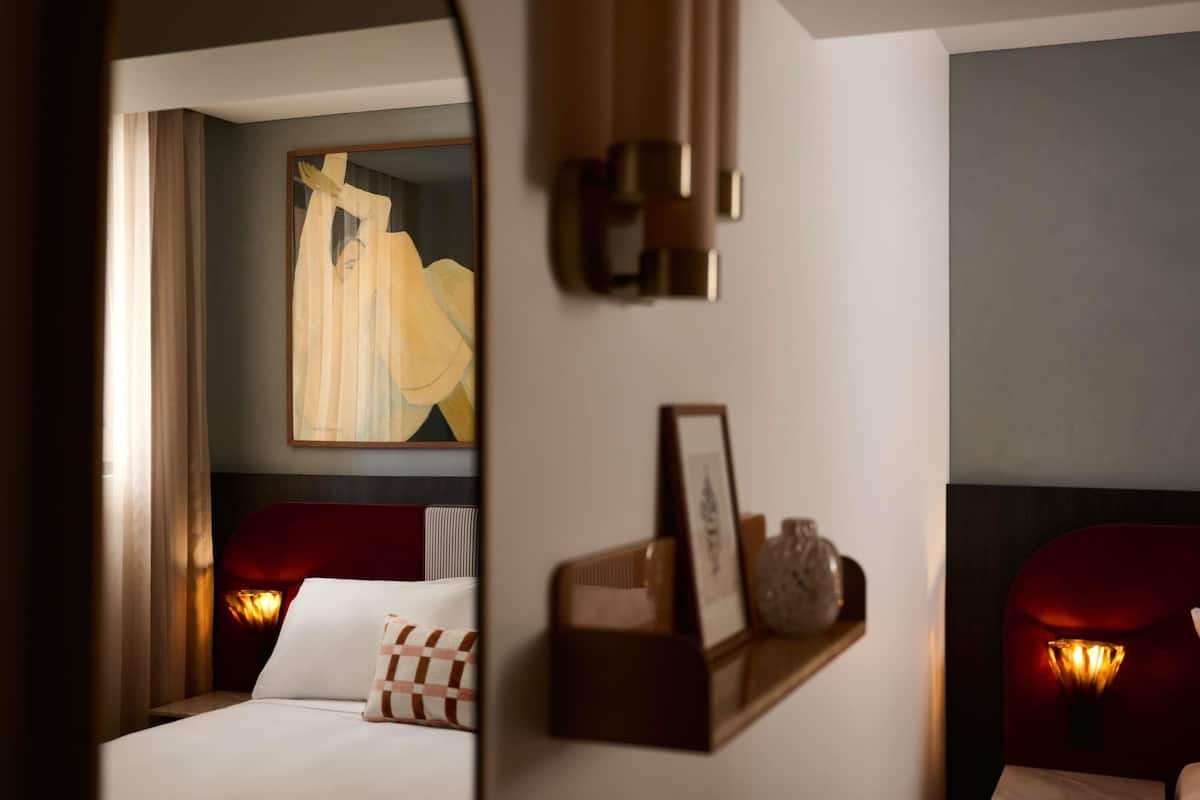 A hotel room with a bed, a nightstand, a wall-mounted shelf with decorative items, and a painting on the wall.