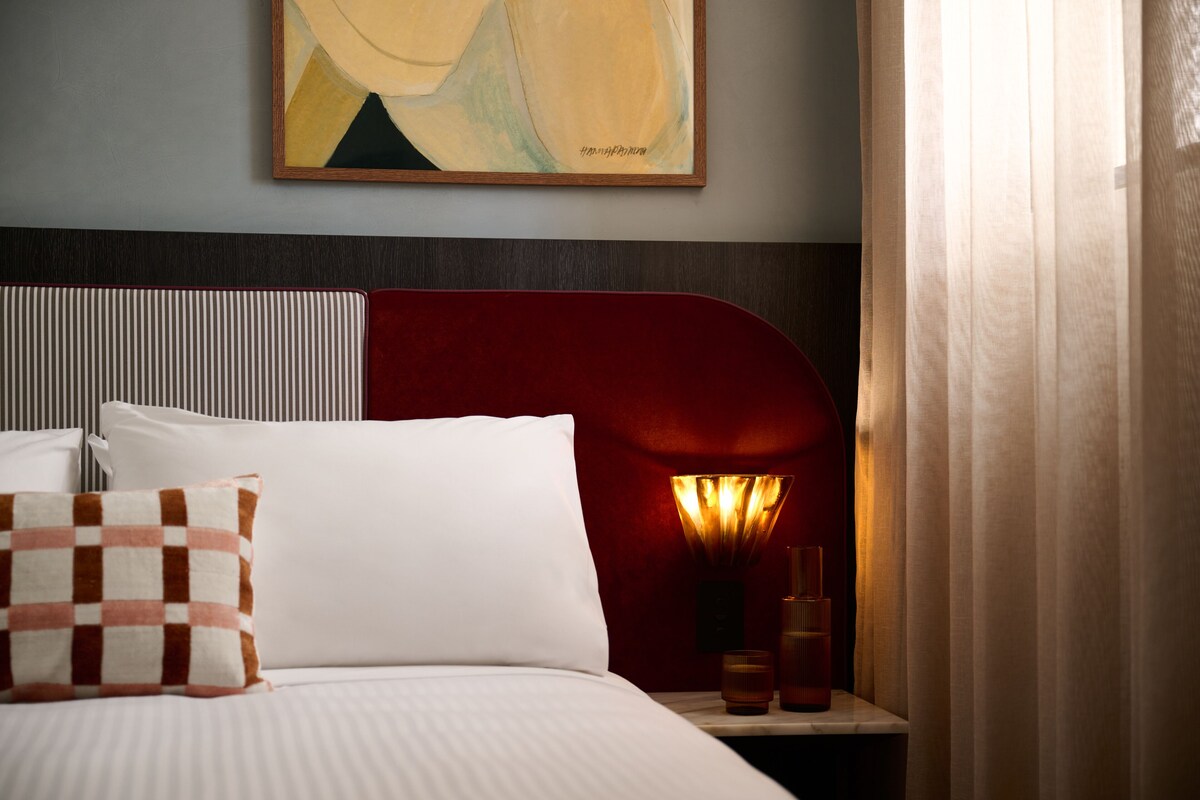Quarto de hotel com cama, almofadas, mesa de cabeceira, abajur e um quadro na parede.