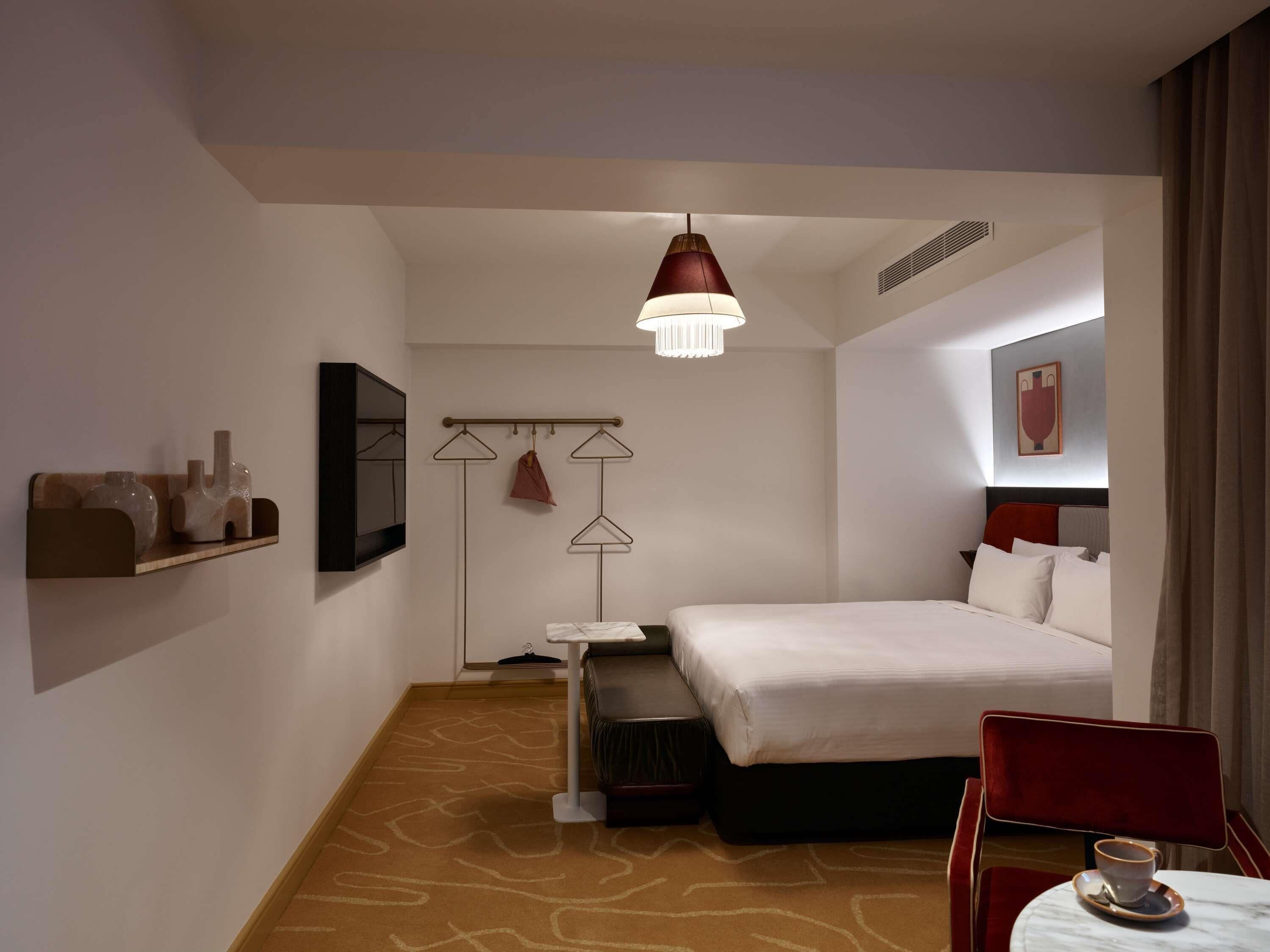 A hotel room with a bed, a small table with a cup, a chair, a hanging light fixture, and a coat rack with clothes.