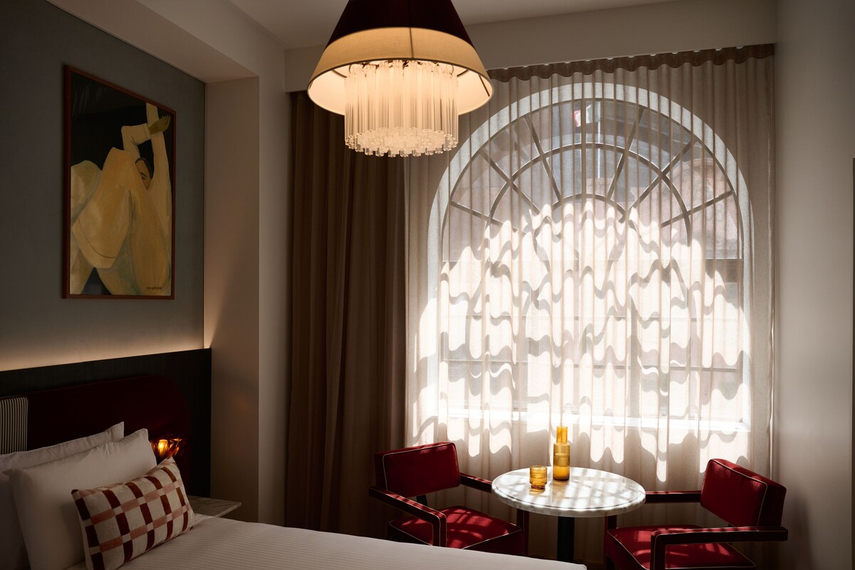 Quarto de hotel com cama, mesa redonda com cadeiras, um lustre e uma grande janela com cortinas.