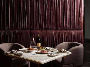 A dining table set with plates, glasses, and a bottle of wine, with a plush, dark-colored upholstered wall behind it. - Hotel Morris Sydney - Handwritten Collection (Haymarket)
