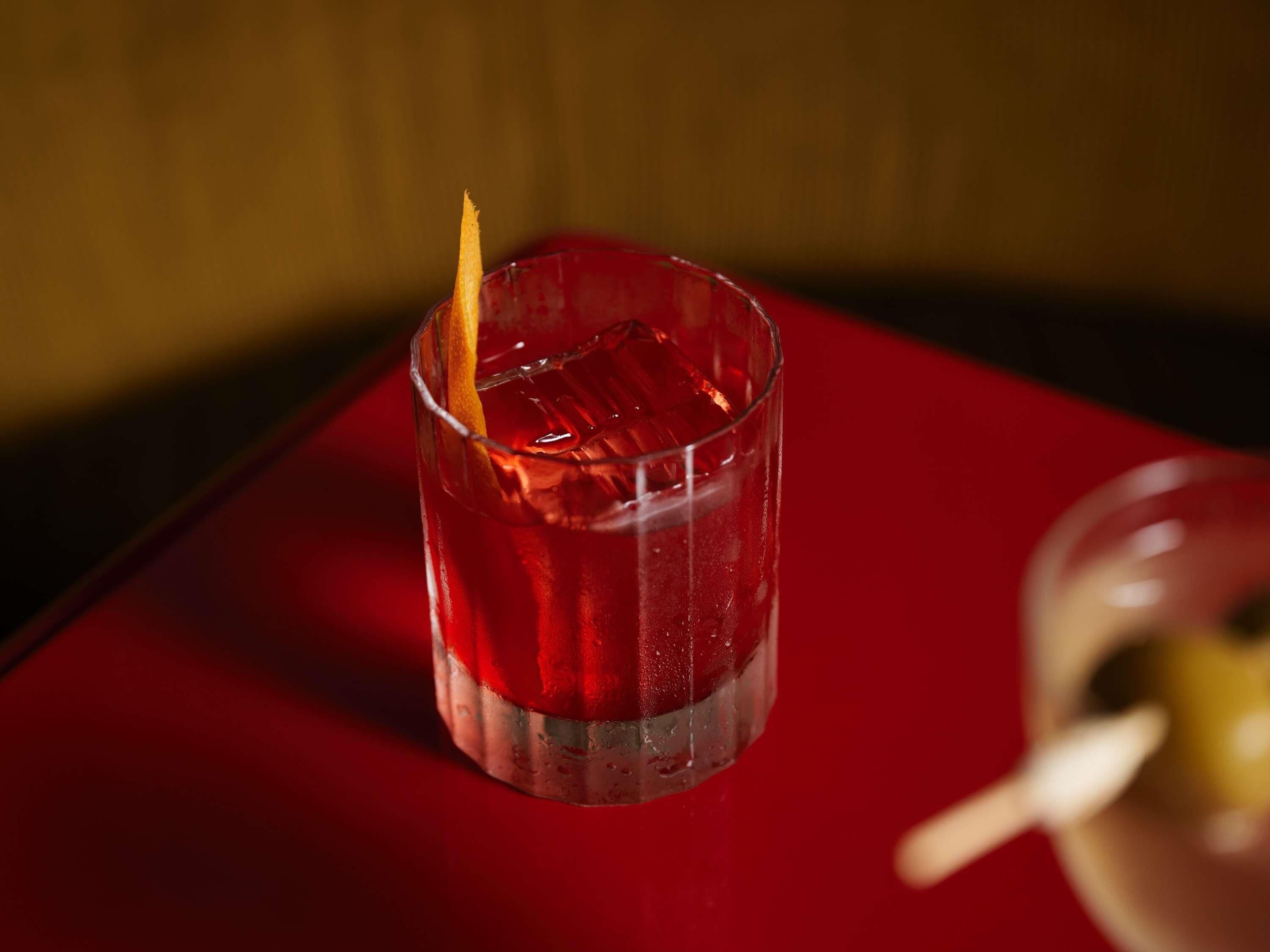 A glass with a red beverage and an orange peel garnish on a red table.