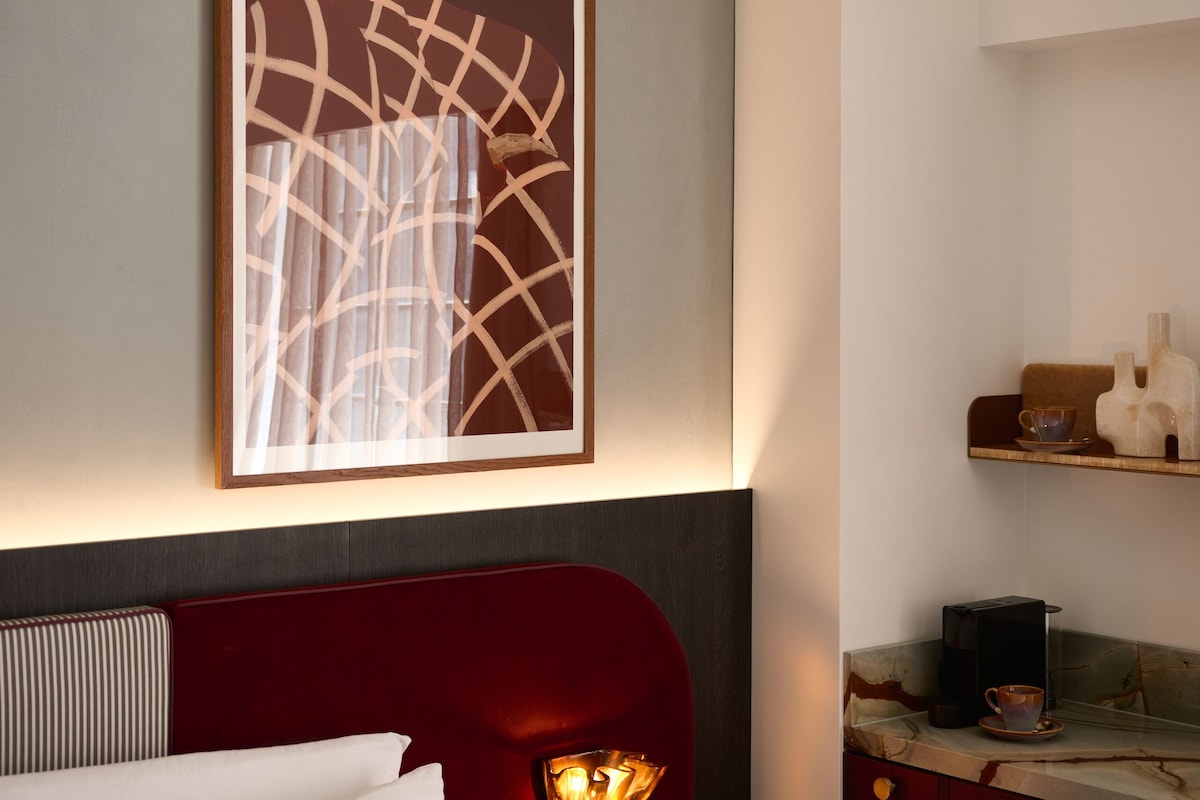 Quarto de hotel com cama, mesa de cabeceira, luminária na parede, obra de arte emoldurada e um pequeno estante com objetos.