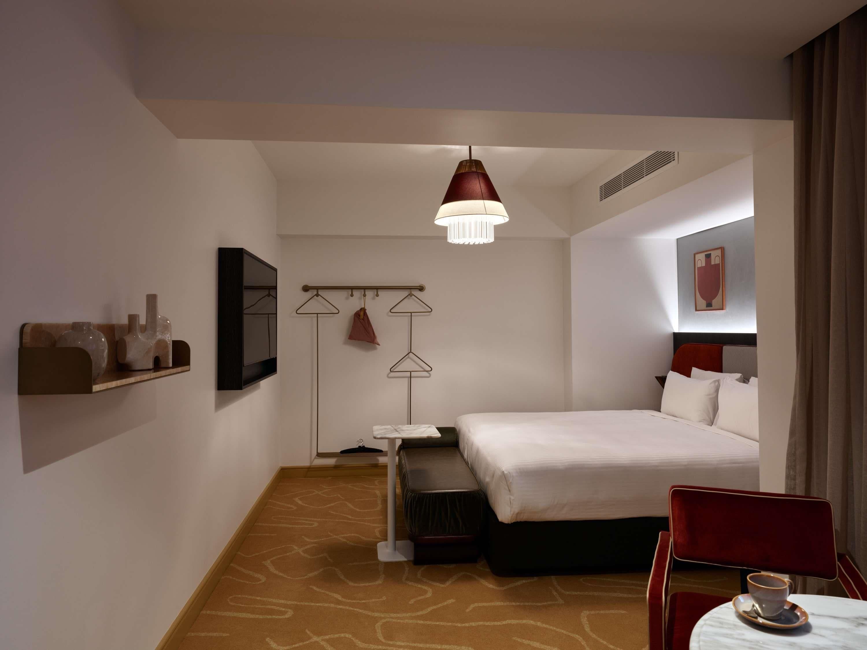 A hotel room with a bed, a small table with a cup, a chair, a hanging light fixture, and a coat rack with clothes.