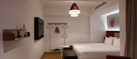 A hotel room with a bed, a small table with a cup, a chair, a hanging light fixture, and a coat rack with clothes.
