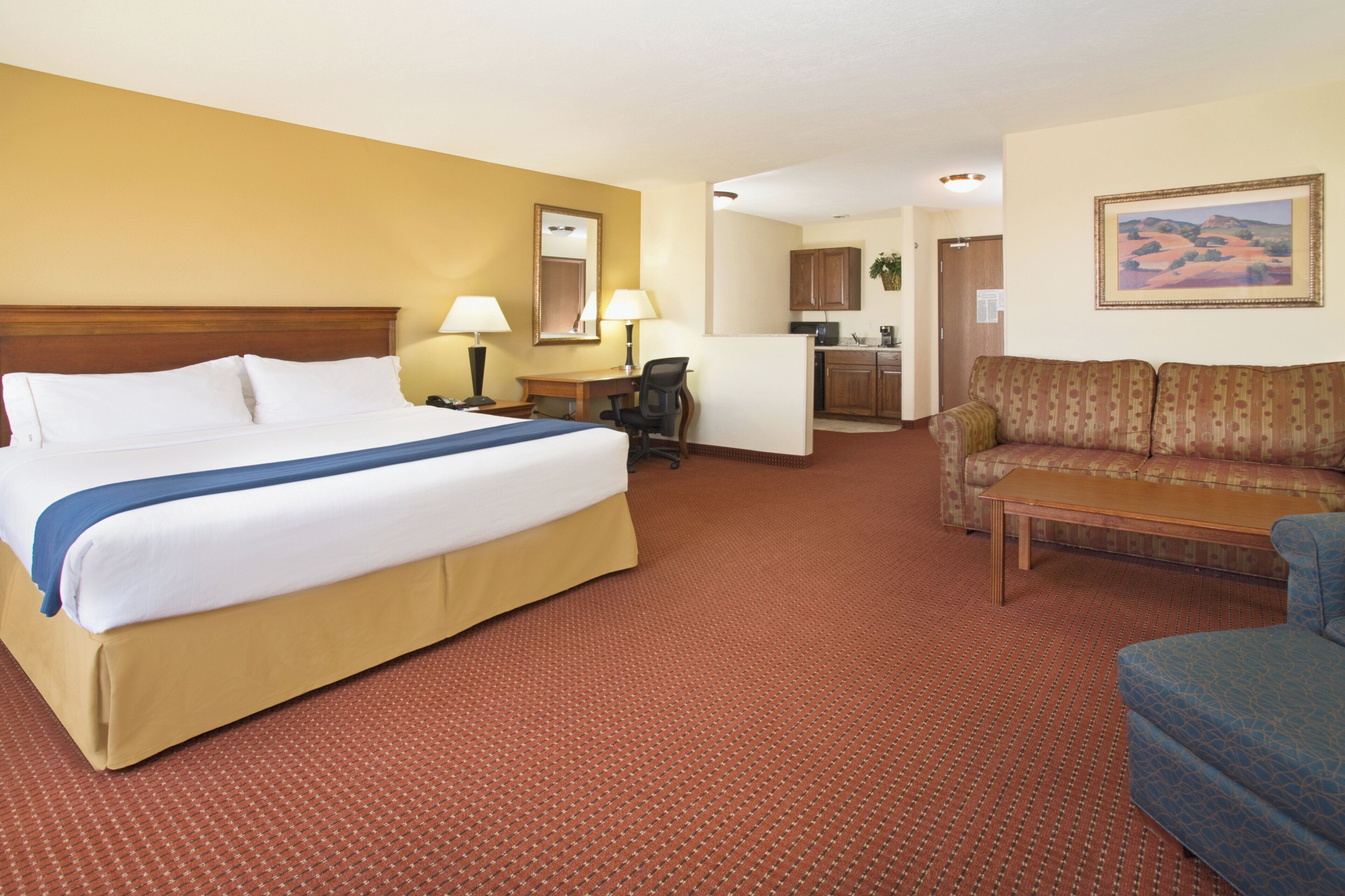Quarto de hotel com uma cama grande, escrivaninha, sofá e cozinha compacta.