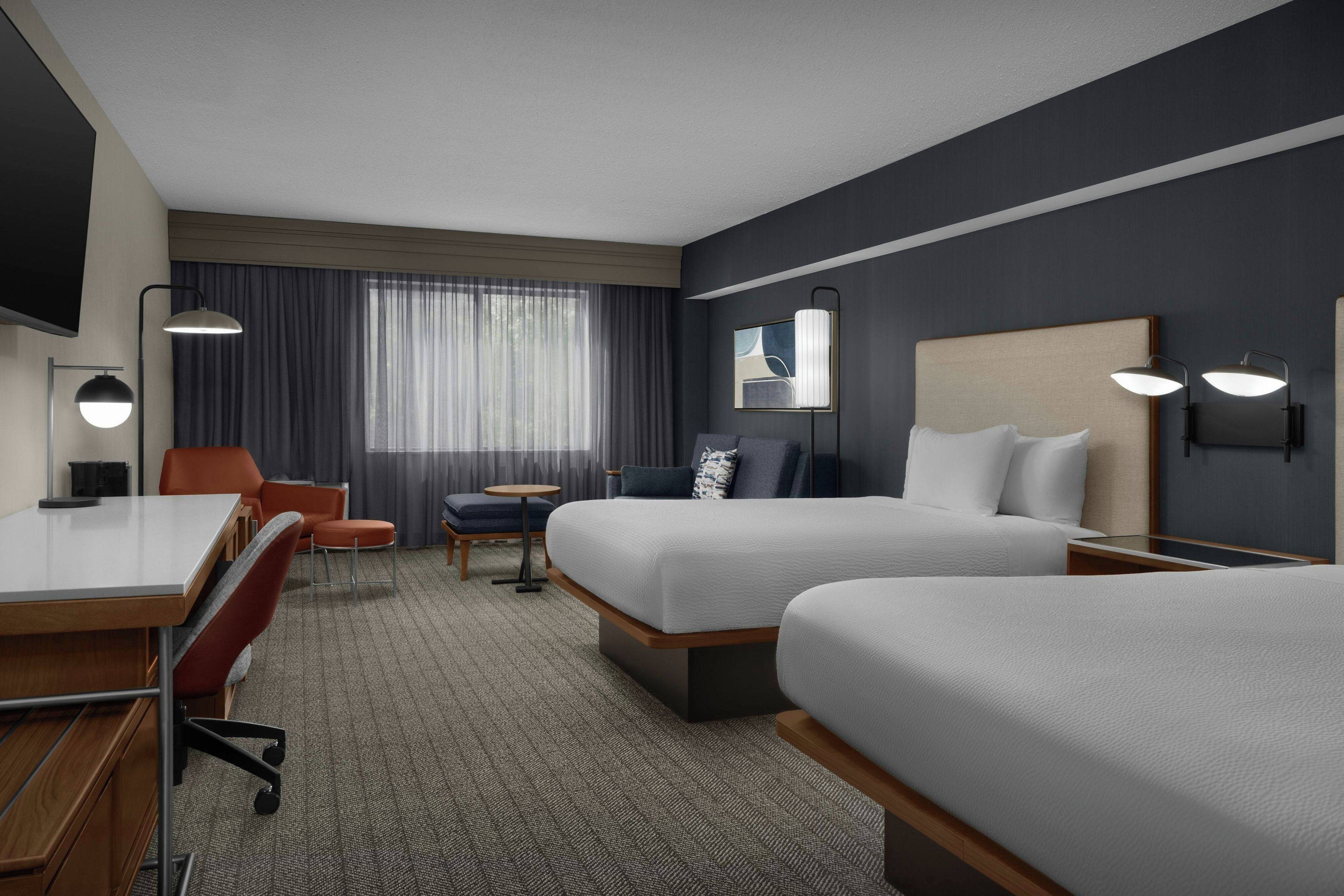 A hotel room with two beds, a desk, a chair, a small table, and a TV.