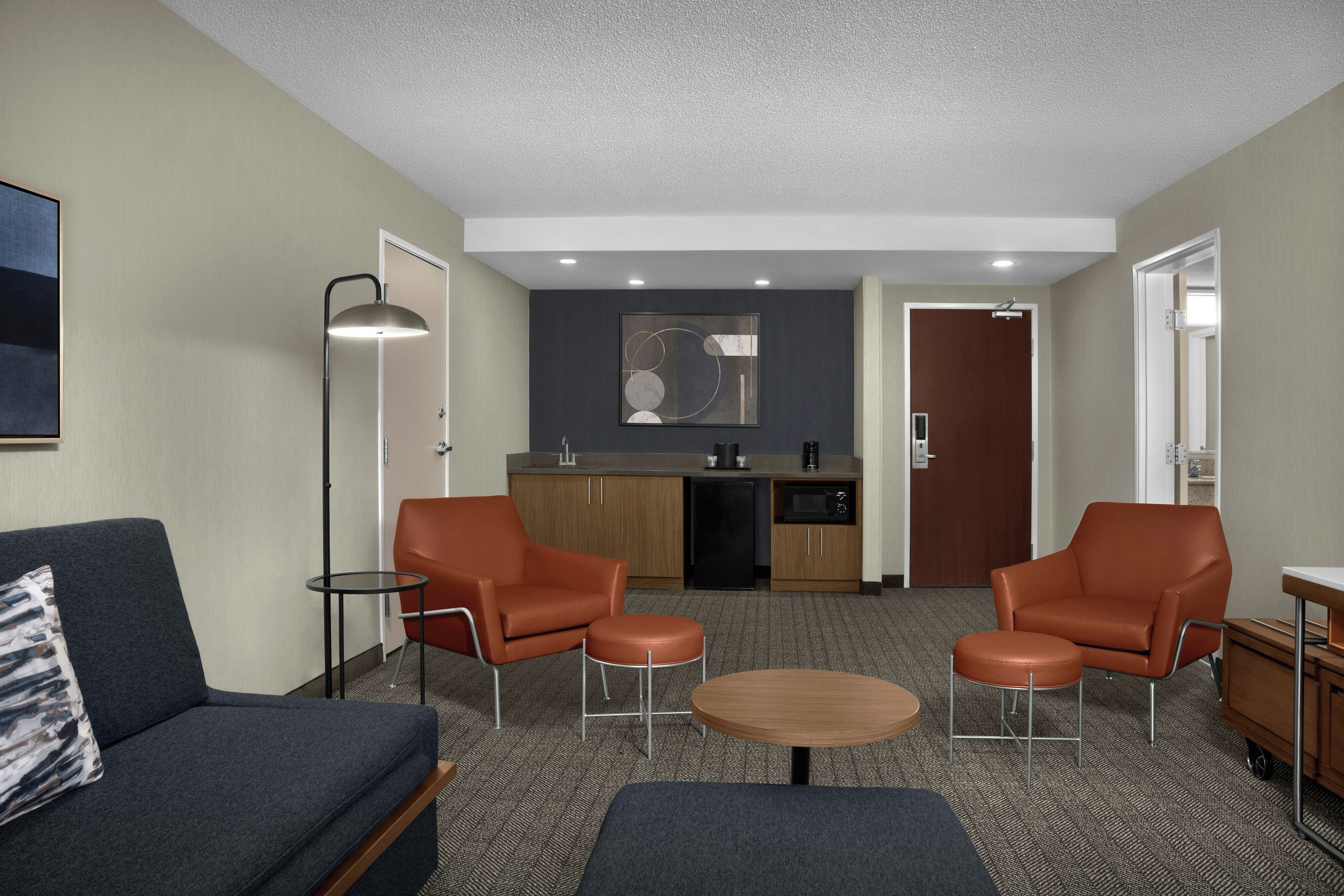 A compact living space with a sofa, two armchairs, a small table, a kitchenette, and a door leading to another room.