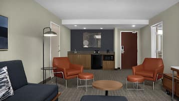 Un espace de vie compact comprenant un canapé, deux fauteuils, une petite table, un coin cuisine et une porte donnant sur une autre pièce.