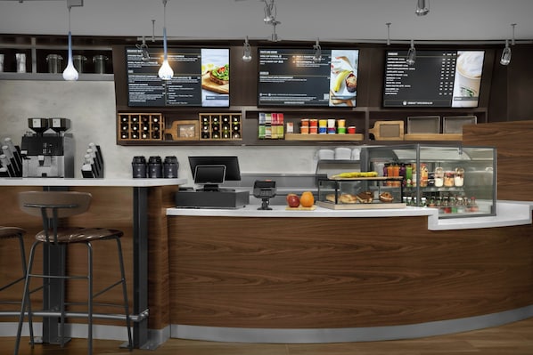 En modern caféinredning med en trädisk, barstolar, en kaffemaskin, en menytavla och ett skyltfönster med olika produkter.