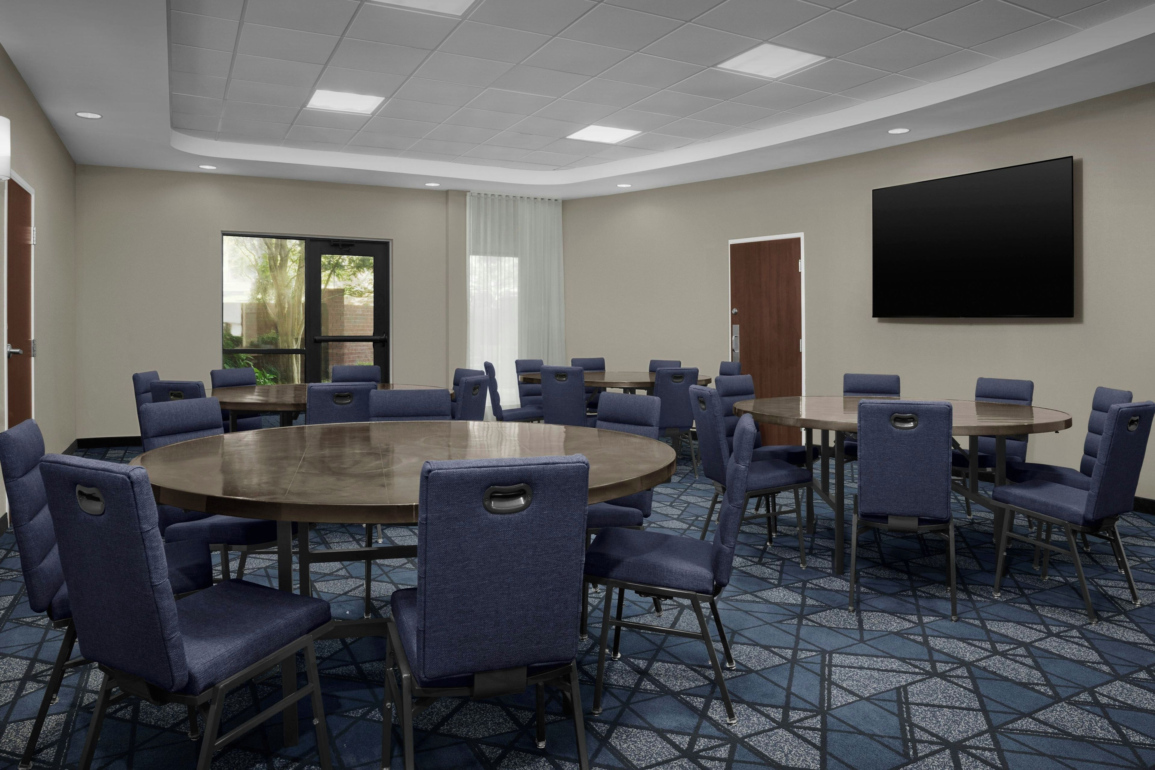 A conference room with round tables, chairs, a large screen, and a view of greenery outside.