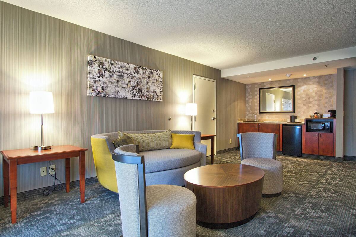 Ein Hotelzimmer mit einer Couch, Sesseln, einem Couchtisch, einem Schreibtisch, einer Lampe, einer Küchenzeile und einem großen Spiegel.