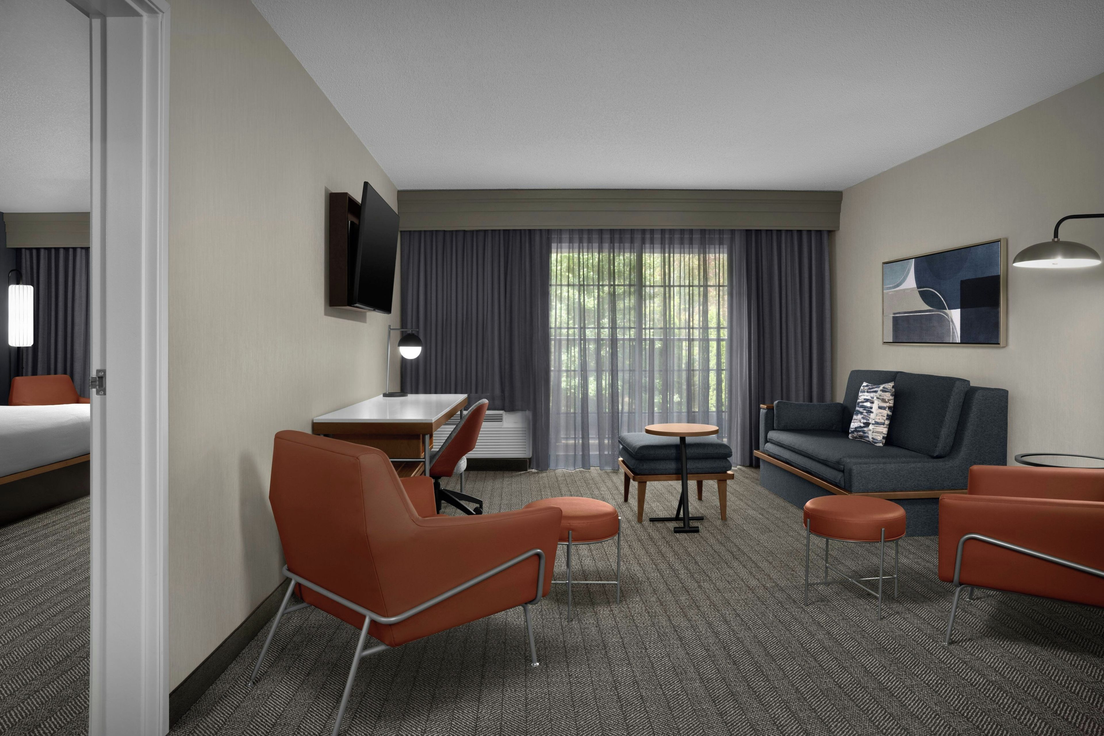 Habitación de hotel con cama, escritorio, sofá, dos sillones, una mesita, televisión montada en la pared y ventana con cortinas.