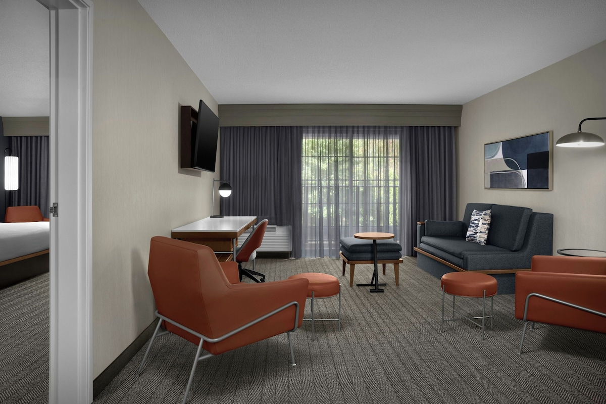 A hotel room with a bed, a desk, a sofa, two armchairs, a small table, a TV mounted on the wall, and a window with curtains.