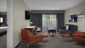 Habitación de hotel con cama, escritorio, sofá, dos sillones, una mesita, televisión montada en la pared y ventana con cortinas.