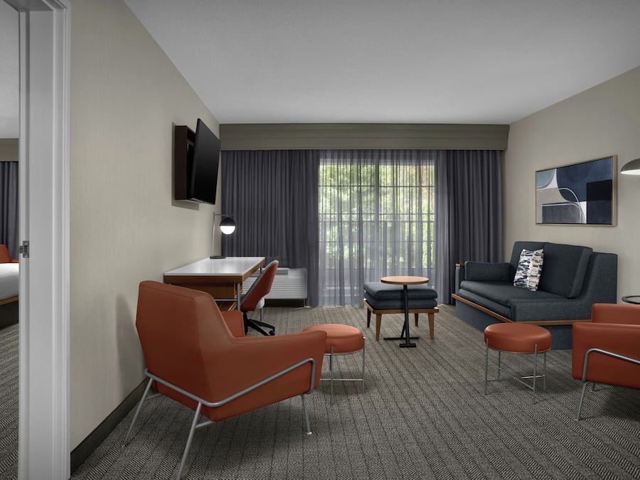 Une chambre d’hôtel comprenant un lit, un bureau, un canapé, deux fauteuils, une petite table, un téléviseur fixé au mur et une fenêtre avec des rideaux.