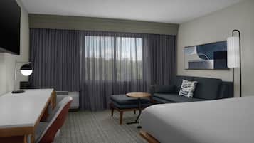 Une chambre d’hôtel comprenant un lit, un bureau, une chaise, une petite table, un canapé, une télévision et une fenêtre avec des rideaux.