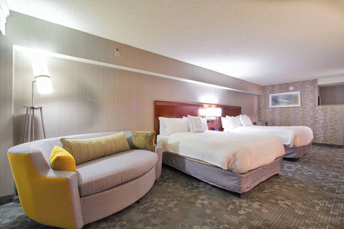 Ein Hotelzimmer mit einer Couch, zwei Betten, einer Lampe und einem an der Wand befestigten Fernseher.