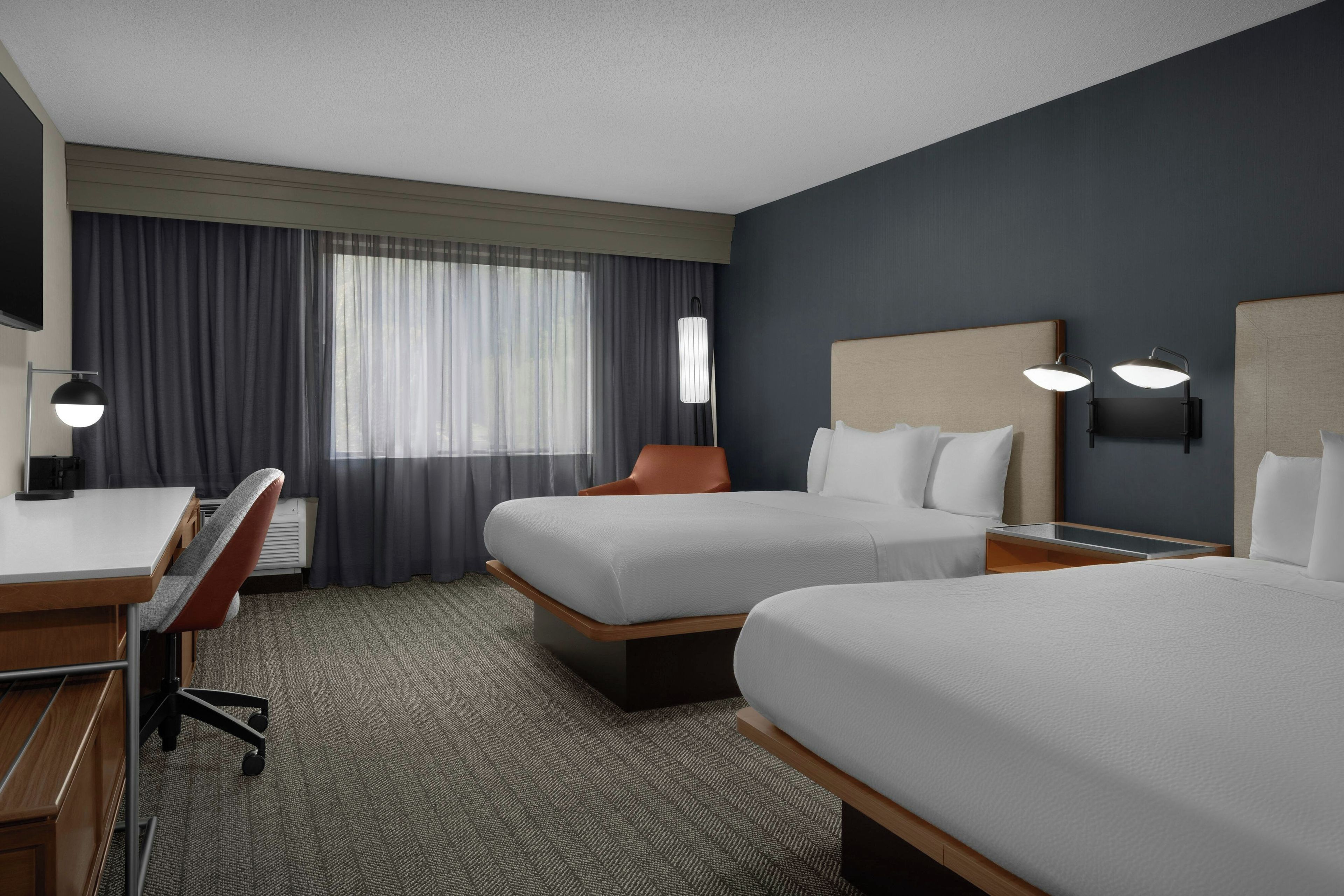 A hotel room with two beds, a desk, a chair, a lamp, and a window with curtains.