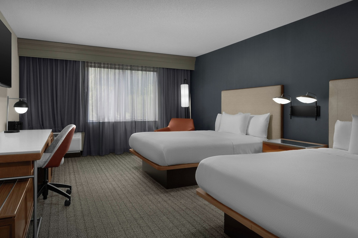 A hotel room with two beds, a desk, a chair, a lamp, and a window with curtains.