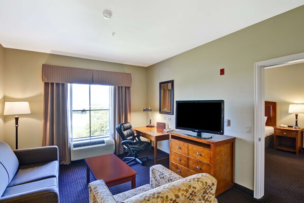 A hotel room with a sofa, a chair, a desk with a television, a dresser, and a window with curtains. - Homewood Suites Houston West Energy Corridor (Houston)