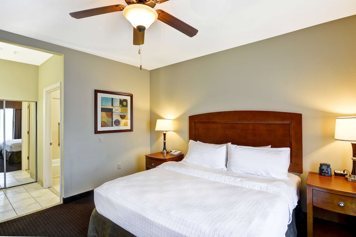 A hotel room with a large bed, a wooden headboard, a ceiling fan, a bedside table with a lamp, and a framed picture on the wall.
